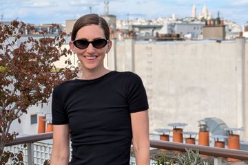 A woman smiling and wearing sunglasses, standing on a balcony with a city view in the background